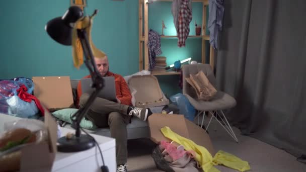 千禧年男人在笔记本电脑前浏览 坐在沙发上 在凌乱的客厅里工作 家里有垃圾 穿着休闲装 复制空间 — 图库视频影像