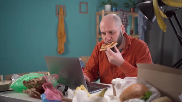Skäggig Man Äter Pizza Och Jobbar Ett Rörigt Rum Med — Stockvideo