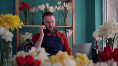 Çiçekçide dizüstü bilgisayarda çalışan yakışıklı küçük işletme sahibi telefonda müşteriyle konuşuyor. Boşluğu kopyala