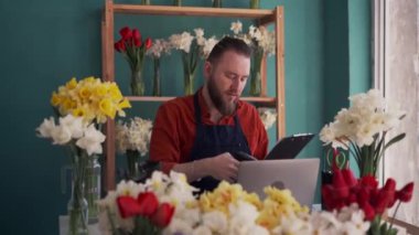 Çiçekçi dükkanının önlük giymiş portre sahibi dizüstü bilgisayarda çalışıyor ve sipariş numaralarını tarıyor. Küçük işletme çiçekçiliği kavramı.