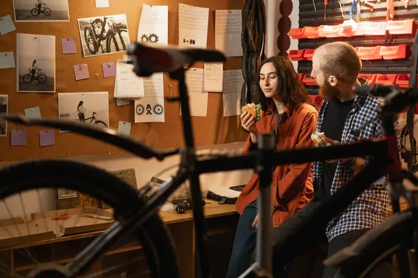 Couple in love chats happily and eats delicious burgers after fixing and servicing a bicycle in repair shop. People working and eating in garage. Copy space