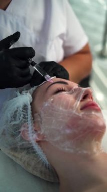 Kozmetik salonunda yaşlanma karşıtı biyo-evyalizasyon. Genç bir kadın için hiyalürik asit derisi alınıyor. Şırıngalı bir güzellik uzmanı. Uzayı kopyala Dikey video