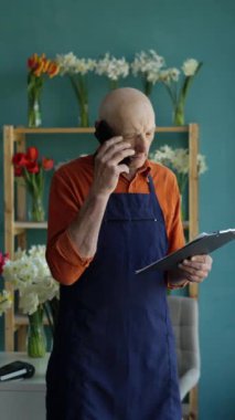 Çiçekçi, yaşlı bir adam akıllı telefondan arıyor ve çiçekçisindeki panoya not alıyor. İnsanlar, perakende, iş ve çiçekçilik konsepti. Dikey video