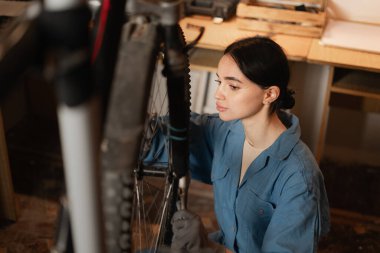 Güzel esmer, uzun saçlı bayan işçi garajda çömelmiş bisikleti tamir ediyor. Motosiklet atölyesinin iç tarafında. Boşluğu kopyala
