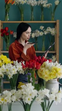 Güzel esmer kız çiçekçi dükkanında çalışırken akıllı telefon kullanarak sesli mesajlar kaydediyor. Boşluğu kopyala