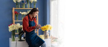 İnsanlar, iş ve çiçekçilik konsepti. Akıllı telefondan arayan ve çiçekçi masasındaki panoya yazı yazan kadın çiçekçi. Pankart. Boşluğu kopyala