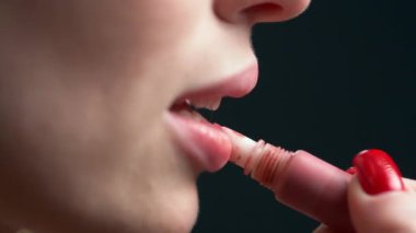 Kırmızı manikürlü bir kadının elinin yan görünüşü dudaklarının üst ve alt dudaklarına ruj sürerken dudaklarını hafifçe açık ve seksi bir şekilde şapırdatıyor..