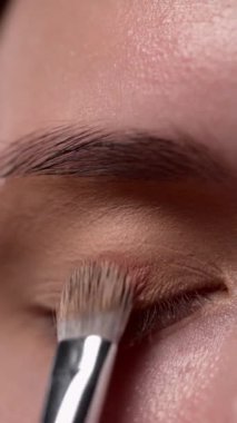 Profesyonel bir makyöz, modelin üst göz kapağına kahverengi göz farı sürmek için fırça kullanır. Yakın plan. Genç beyaz kadın.