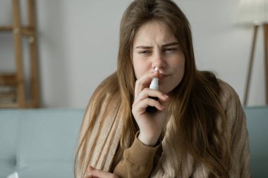 Soğuk algınlığıyla mücadele eden, ev ortamında battaniyenin altında kanepede oturan beyaz bir kız tıbbi ürünler uyguluyor ve burun akıntısını tedavi etmek için burun spreyi kullanıyor..