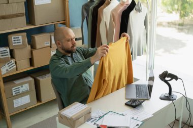 Yorgun bir adam müşterisinin elbisesini elinde tutuyor. Bilgisayardaki çevrimiçi sırayla kontrol ediyor. Boşluğu kopyala