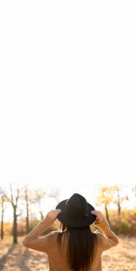 Sonbahar ormanında durup gökyüzüne bakan bej şapkalı bir kadının arka görüntüsü. Özgürlük ve doğa kavramı. Dikey pankart