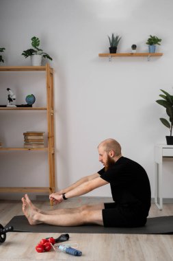 Bir adam evinde yoga yapıyor. Etrafı bitki ve raflarla çevrili. Huzurlu bir atmosfer yaratıyor.