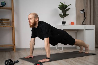 Bir adam, kapalı bir yoga minderinin üzerinde, bir ev ortamında sağlık ve refah üzerine odaklanarak kalas pozu veriyor.