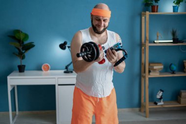 Turuncu saç bandı ve şort takan neşeli bir adam, dambılı kaldırıyor ve mavi arka planla evde çalışıyor..