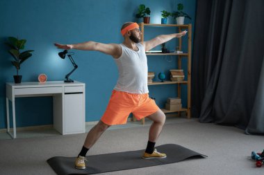 Turuncu şort ve saç bandı takan bir adam oturma odasında yoga yapıyor, yoga minderinde Warrior Poz çalışıyor.
