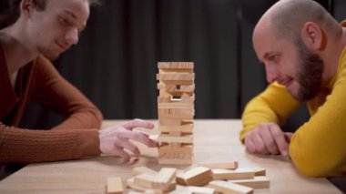 İki adam dikkatle Jenga oynuyor, dikkatle tahta blokları tutarken konsantrasyon ve stratejik hareketler sergiliyorlar. Boşluğu kopyala