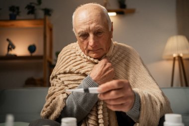Battaniyeli sağlıksız bir adam sıcaklığını termometreyle ölçer. Soğukkanlı, tedavi edilecek. Boşluğu kopyala