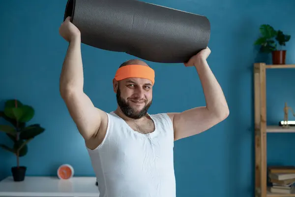 stock image A man smiles, holding a yoga mat over his head, ready for home workout, showing motivation and dedication to fitness