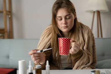 sick woman wrapped in blanket holds thermometer in one hand and cup of tea in the other. Treatment at home. Taking medications. copy space clipart