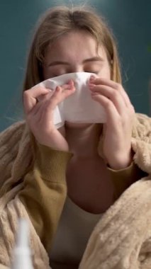 Genç bir kadın hastalandı. Battaniyeli kadın evinde kanepeye oturdu, burnunu peçeteyle sildi. Dikey video. Yakın plan.