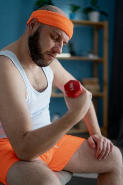 Sakallı, tişörtlü ve şortlu komik adam evde küçük dambıllarla egzersiz yapıyor. Boşluğu kopyala