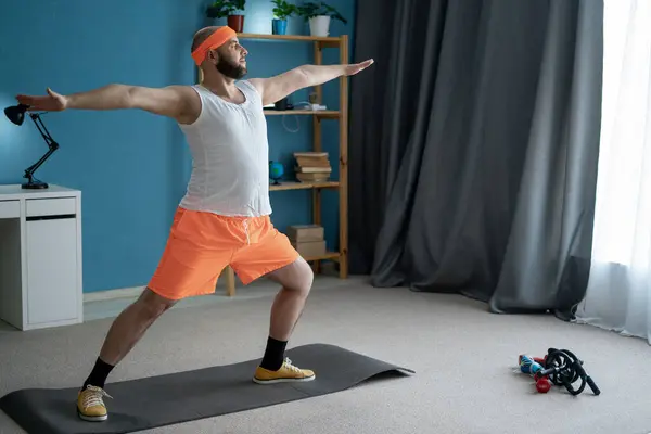 stock image A man with a beard is practicing yoga at his home, wearing orange shorts and a headband, executing a warrior pose