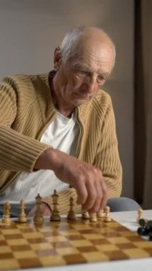 Yaşlı bir adam stratejik bir satranç maçına dalmış, evindeki tahtanın üzerindeki taşları dikkatlice hareket ettiriyor.