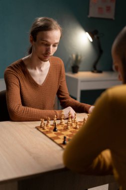 Kahverengi kazaklı genç bir adam, bir satranç maçına odaklanmış, ahşap bir masada oturuyor ve rakibi samimi bir iç mekanda oturuyor. Bir masa lambası arka planı aydınlatır