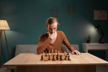 Kahverengi kazaklı genç adam bir sonraki hamlesini bir satranç oyununda ahşap masada oturarak düşünüyor. Odada bir masa lambasından gelen deniz mavisi duvarlar ve sıcak ışıklandırma bulunuyor..