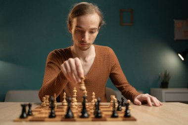 Kahverengi kazaklı genç adam bir satranç oyunu oynamaya, ahşap masada oturmaya derinden konsantre oldu. Oyunun odak noktası konsantrasyon ve stratejik düşünce..