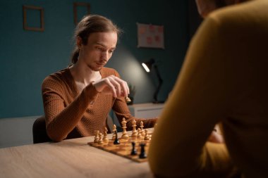 Kahverengi kazaklı genç adam ahşap bir masada otururken satranç stratejisini planlıyor. Rakibi, kısmen görünür, sarı bir kazak giyiyor..