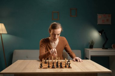 Kahverengi kazaklı genç bir adam, bir satranç oyununda, ahşap bir masada oturuyor. Odada bir masa lambasından gelen deniz mavisi duvarlar ve sıcak ışıklandırma bulunuyor..