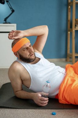 Orta yaşlı sporcu yoga minderinde oturuyor ve spor salonunda su içiyor. Boşluğu kopyala