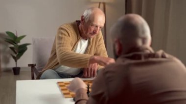 Yaşlı bir adam arkadaşlarıyla samimi, parlak bir odada satranç oynar, zihinsel uyarılma ve sosyal etkileşimden zevk alır.