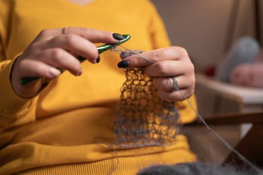 Gri iplik örmek için örgü kancası kullanan ellerin yakın çekimi. Eller dikkatle bir dikiş oluşturuyor, bu da zanaatın kesin hareketlerini gösteriyor..