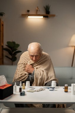 Yaşlı sağlıksız bir adam, battaniyeye sarılmış ve hasta bir şekilde kanepede oturuyor. Hastalığın ilacı gözünün önündeydi. Boşluğu kopyala