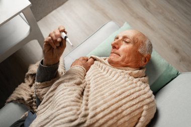 Yaşlı bir adam kanepede yatar, termometreyle ateşini ölçer, battaniyeye sarınır, hastalığını ve iyileşmesini evde tek başına idare eder..
