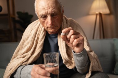 Yaşlı bir adam kanepeye oturur, battaniyeye sarınır, elinde bir hap ve bir bardak su, kendini kötü hisseder ve ilaçlarını almaya hazırlanır..