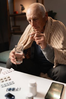 Yaşlı bir adam kanepeye oturur, battaniyeye sarınır, elinde bir hap ve bir bardak su, çeşitli ilaçlarla sarılı, kendini kötü hisseder ve ilacını almaya hazırlanır..