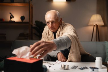 Yaşlı bir adam kendini iyi hissetmiyor, battaniyeye sarılmış peçetelere uzanıyor. Masanın üzerinde çeşitli ilaçlarla çevrili. Bu da hastalığını evde tek başına tedavi ettiğini gösteriyor..