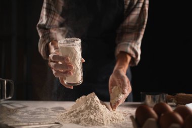 Ekose gömlekli fırıncı hamur hazırlıyor, bardaktan unu masanın üstüne döküyor. Elleri un kaplı, arka planda kaşık, yağ ve yumurta gibi mutfak aletleri var..