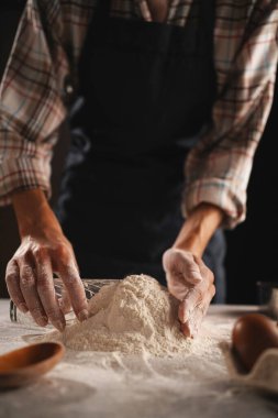 Ekose gömlekli fırıncı masanın üzerindeki bir bardağa un topluyor. Elleri un kaplı ve mutfak aletleri arka planda tahta kaşık ve yumurta gibi. Karanlık, karamsar ışıklandırma sahneyi güçlendirir.
