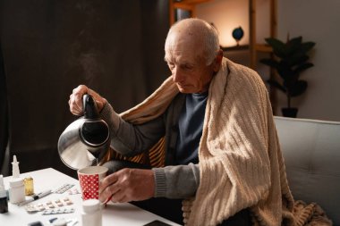 Etrafı ilaçlarla çevrili, battaniyeye sarılmış sıcak çay, ev hastalığı yönetimi ve kendine bakım kavramı olan yaşlı bir adam. Boşluğu kopyala