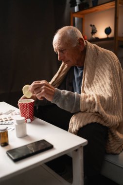 Sıcak bir battaniyeye sarılmış emekli, çayına bir dilim limon ekleyerek kırmızı bir benekli fincana sarılmış. İlaçlar masanın her tarafına yayılmış. Sıcak ev bakımı, onun hastalık sırasında sağlığına dikkat etme çabalarını vurguluyor.
