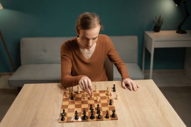 Evde satranç oynayan, rahat bir iç mekanda, ahşap bir masada oturan odaklanmış bir yüzü olan genç adam. Satranç tahtasındaki bir sonraki stratejik hamlesini tasarlarken atmosfer...