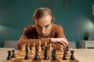 Genç adam bir sonraki hamlesini planlamak için yoğun bir şekilde bir satranç oyununa odaklanmış. Rahat bir ev ortamında oynuyor, stratejik becerilerini geliştirirken entellektüel hobisinin tadını çıkarıyor. Boşluğu kopyala