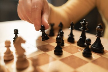 Stratejik bir oyun sırasında tahta bir satranç tahtasına hamle yapan bir elin yakın plan görüntüsü. Sahne satranç taşlarına ve oyuncunun eline odaklanıyor. Oyunun şiddetini vurguluyor.