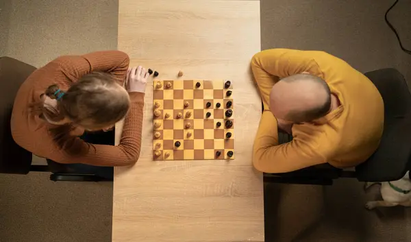 stock image Overhead view of two men playing chess at home. Both players are focused on the game, displaying strategic thinking. The wooden chessboard is set on a light wooden table in a cozy, indoor environment