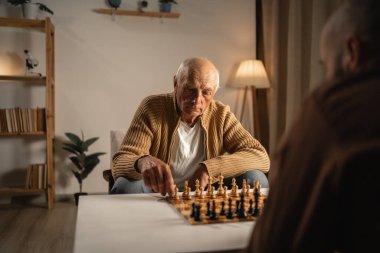 Yaşlı bir baba ve yetişkin oğlu evde satranç oynuyorlar. Oğlu izlerken baba hamle yapıyor. Aile bağları, stratejik düşünme ve kapalı alandaki faaliyetler kavramı.