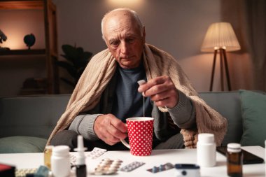 Yaşlı adam battaniyeye sarılmış, sıcak çay hazırlıyor, ilaçlarla çevrili, soğuk algınlığı ve grip için ev tedavisini gösteriyor. Boşluğu kopyala